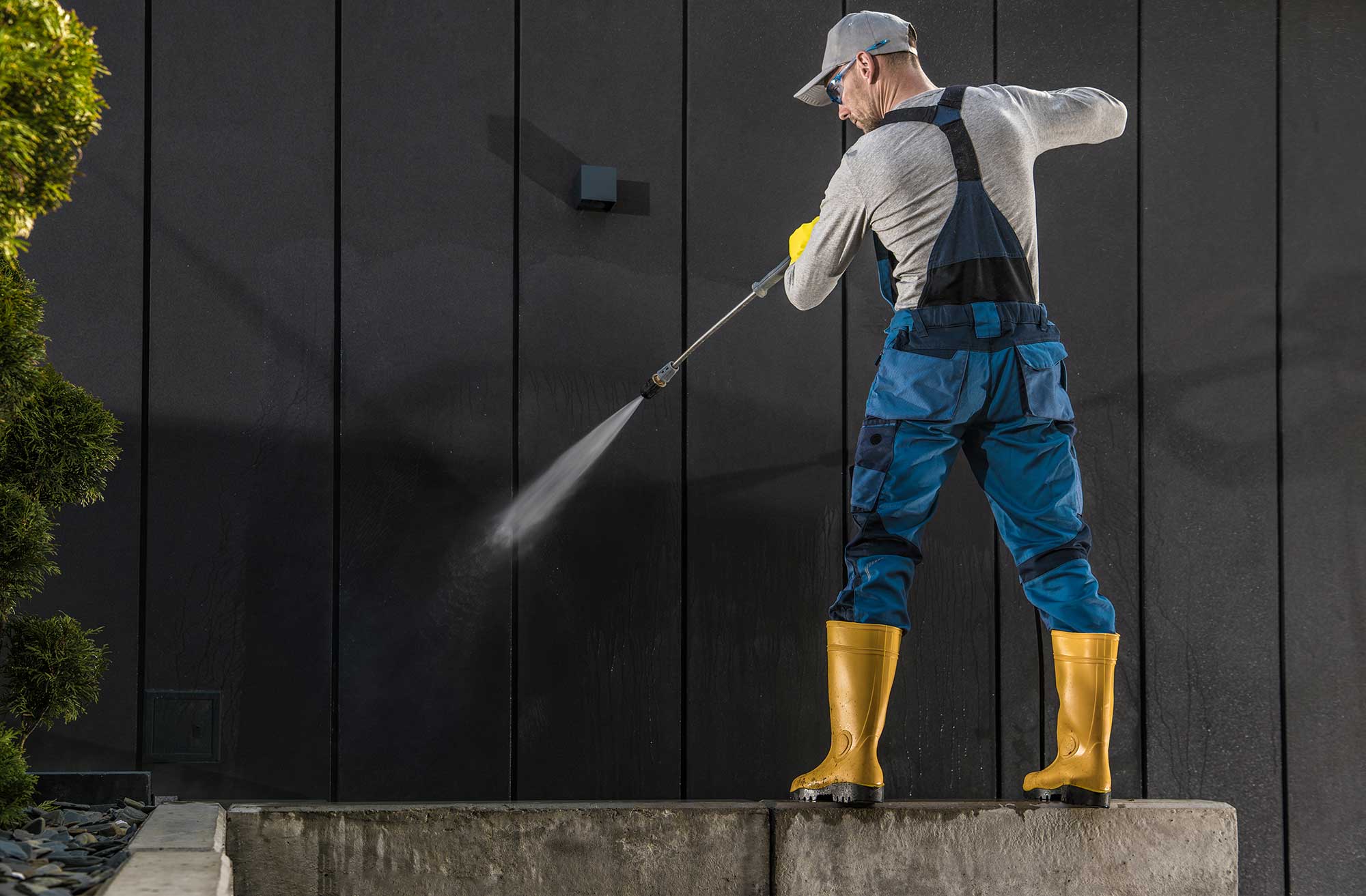 A man in work overalls and yellow boots uses a pressure washer on a dark wall, conveying a sense of cleanliness and industriousness.