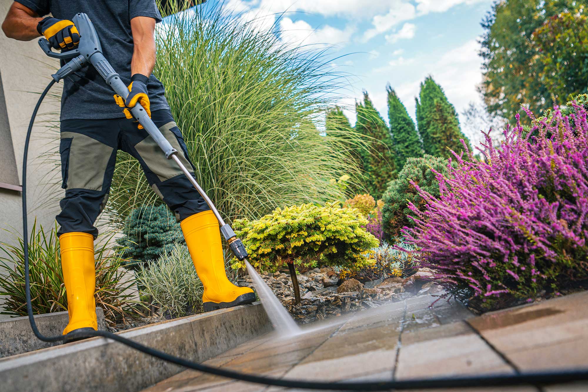 A person in yellow boots uses a pressure washer to clean a patio surrounded by vibrant green and purple foliage, under a blue sky with clouds.