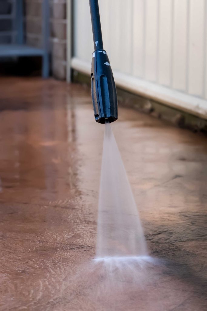 A pressure washer sprays a strong jet of water on a brown, stained concrete surface near a white wall, effectively cleaning the area.